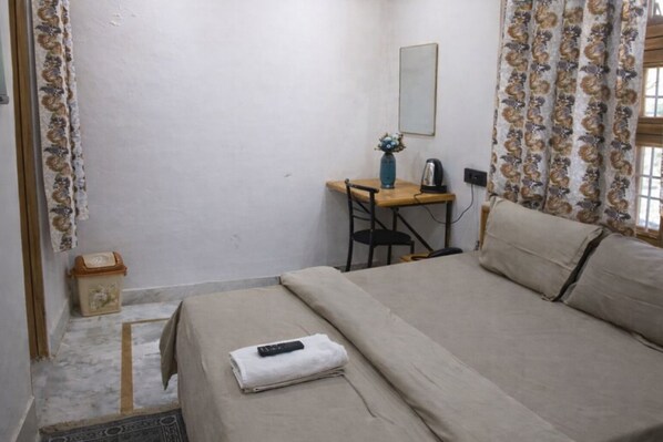 Une petite pièce simple, avec un lit, un bureau et une chaise. Il y a un miroir au mur, une fenêtre avec des rideaux et une petite poubelle.