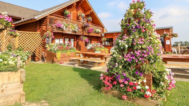 A wooden log cabin with balconies adorned with flowers, a grassy yard, and a wooden fence. - Little Bears Hideaway @ 23 Lakeside (Morpeth)