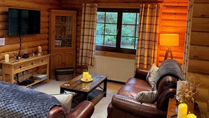 A cozy log cabin interior with a leather sofa, a wooden table with candles, a television mounted on the wall, and plaid curtains. - Little Bears Hideaway @ 23 Lakeside (Morpeth)