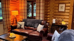 A cozy log cabin interior with a leather sofa, plaid curtains, a lamp, and a small table with candles. - Little Bears Hideaway @ 23 Lakeside (Morpeth)