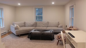 A living room with a beige sofa, a small ottoman, a white desk, and a window with blinds. - 2BR/2BA in a Duplex; Private entry & free parking; Downtown Holliston (Holliston)