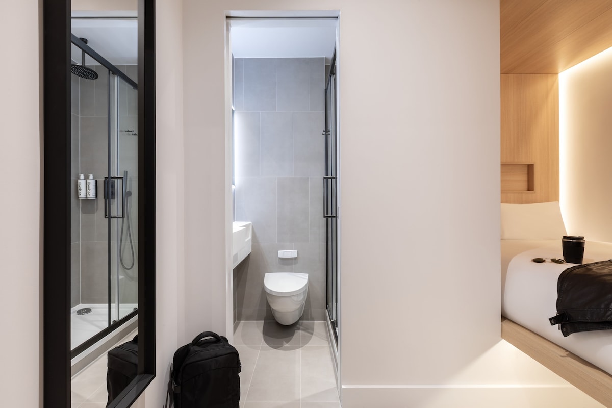 Een moderne badkamer met een glazen douche, een toilet en een bed met nachtkastje.