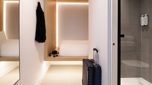 A modern hotel room with a bed, a small seating area, a suitcase, and a shower area.