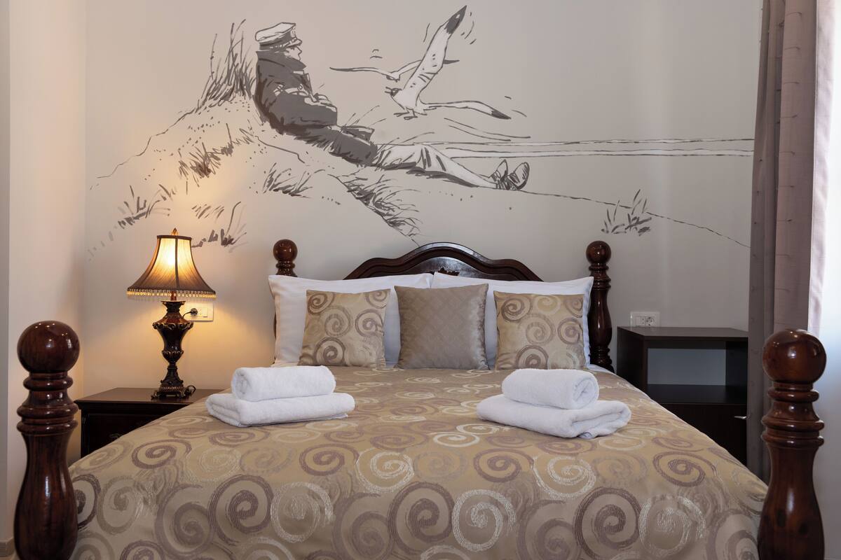 Un llit ben arropat amb un estovat estampat, un mural a la paret, una làmpada de nit i una taula de nit de fusta.