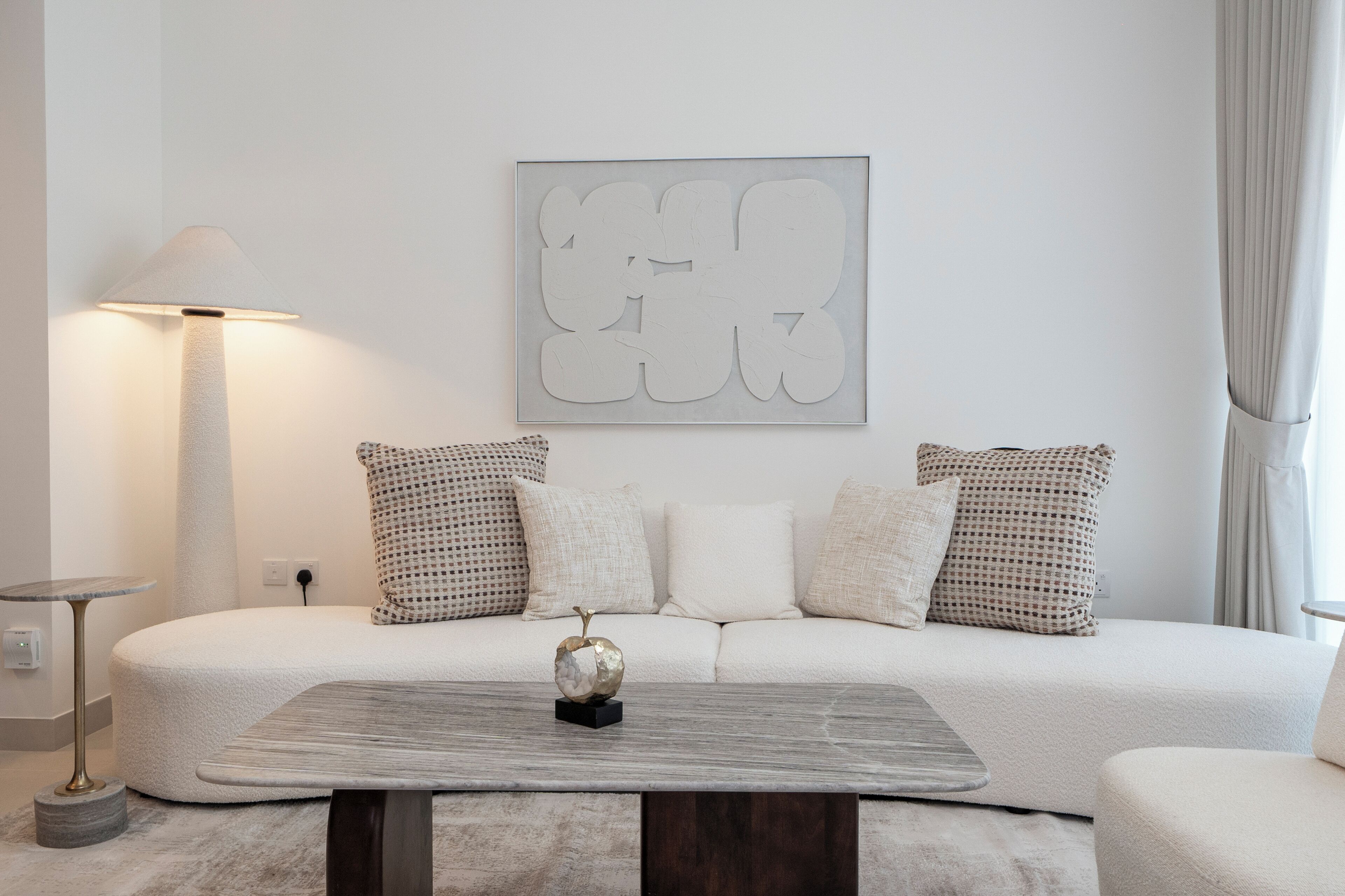Ein modernes Wohnzimmer mit einem weißen Sofa, gemusterten Kissen, einem runden Couchtisch mit einem dekorativen Gegenstand und einem abstraktem Kunstwerk an der Wand.