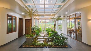 A hotel lobby with a glass ceiling, potted plants, a stone pathway, and a dining area visible through glass doors. - Pataco Tam Coc Hotel (Nam Hoa Lu)
