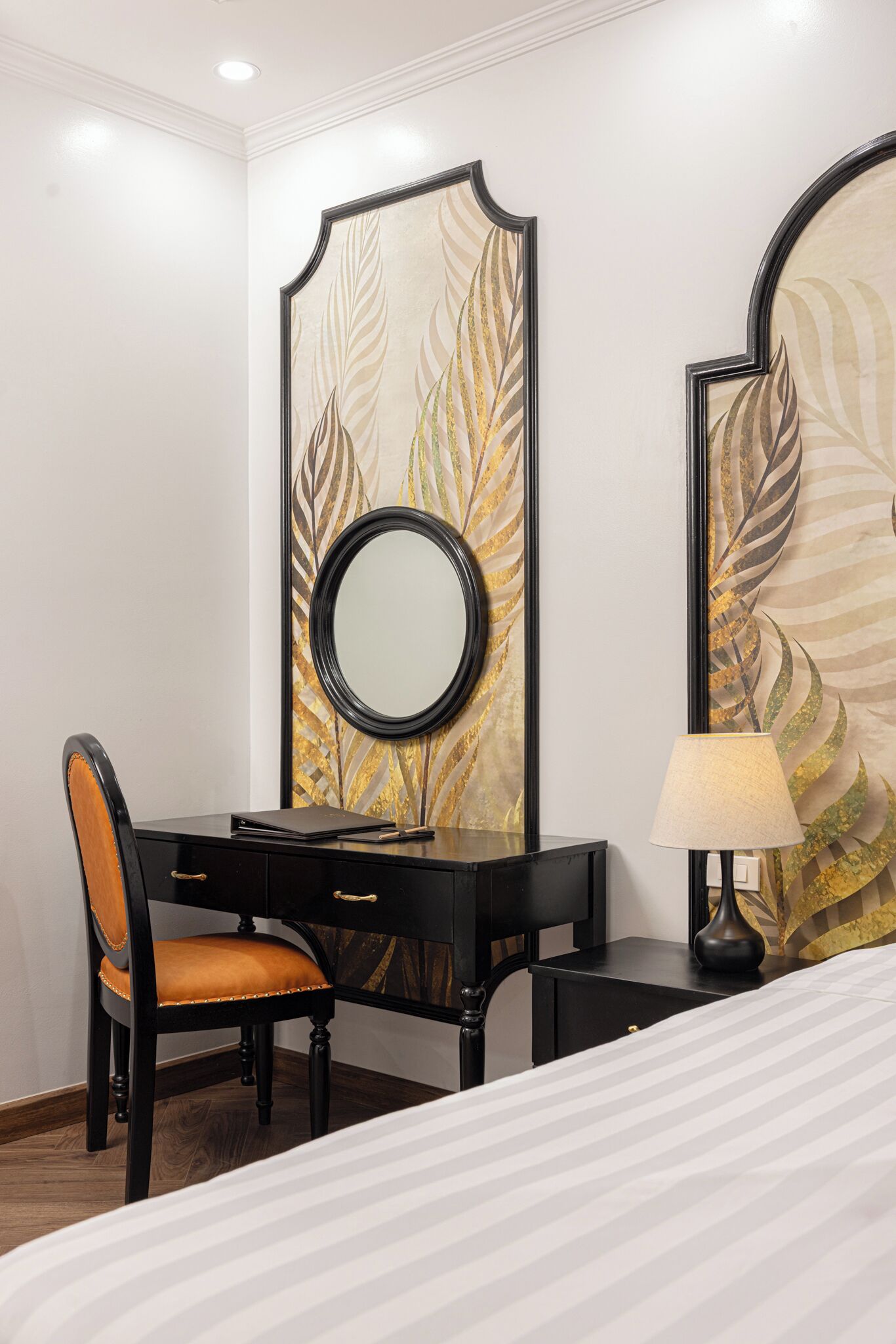 A hotel room with a bed, a desk with a chair, a lamp, and a large mirror with a leaf pattern.