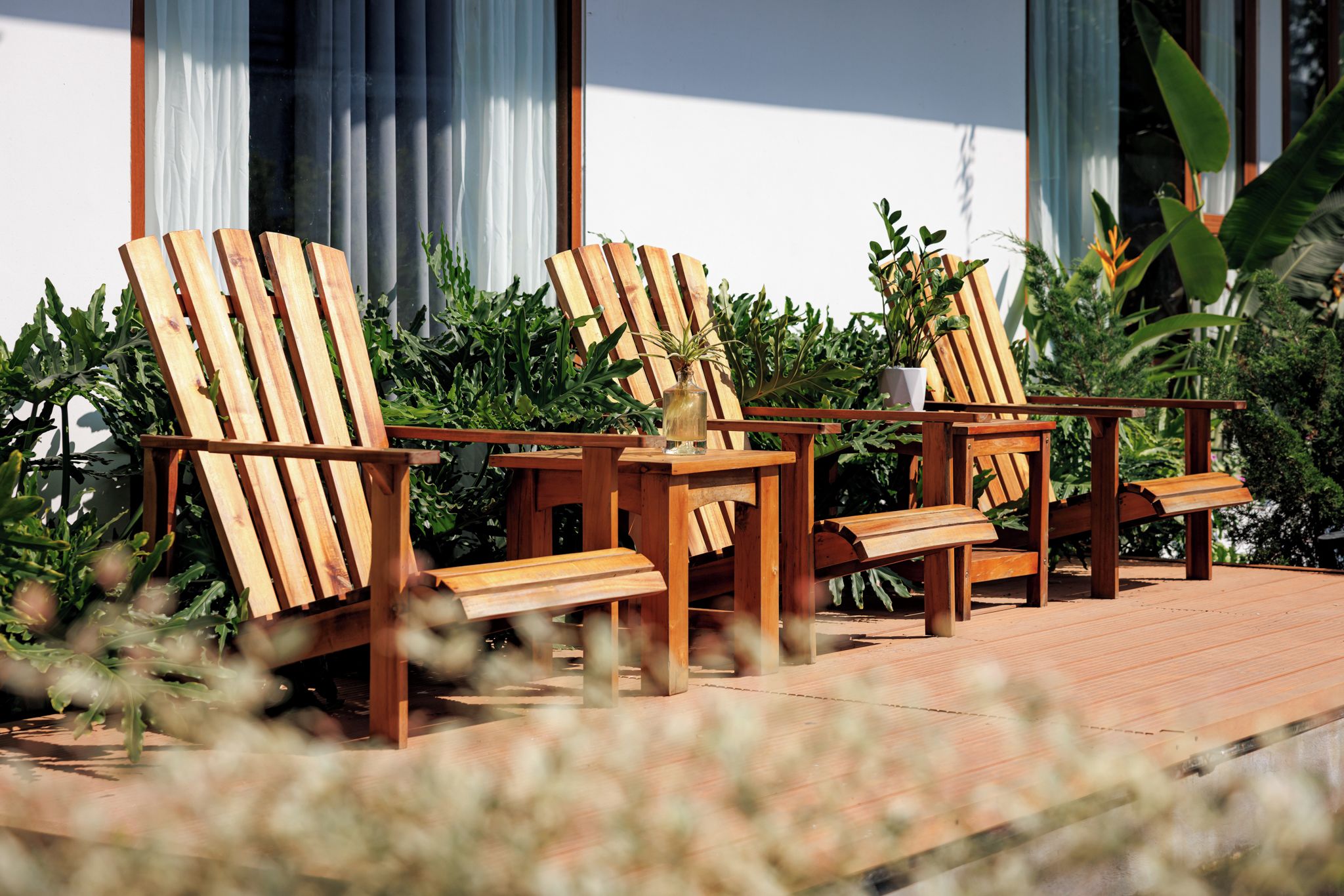 A wooden deck with two lounge chairs and a small table, surrounded by greenery and a building with large windows.