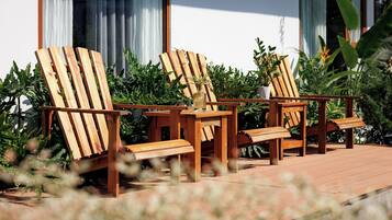 A wooden deck with two lounge chairs and a small table, surrounded by greenery and a building with large windows.