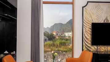 Een moderne woonkamer met twee oranje fauteuils, een ronde glazen tafel, een zwarte kast en een groot raam met uitzicht op bergen en gebouwen.