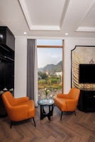 Una sala de estar moderna con dos sillones naranja, una mesa redonda de vidrio, un mueble negro y un ventanal con vistas a montañas y edificios.