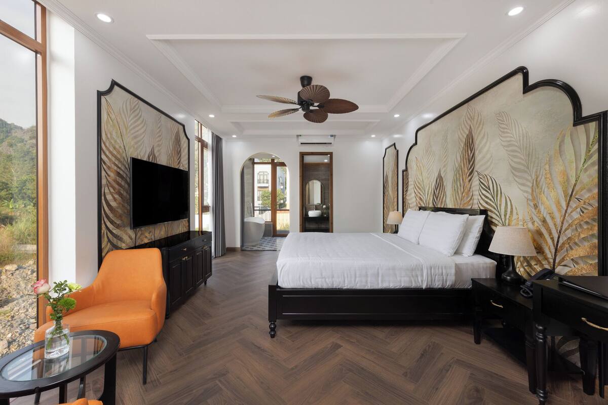 A hotel room with a large bed, a TV, an orange chair, and a view of the outdoors.