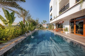 Une piscine dotée d’un bassin aux eaux d’un bleu cristallin, entourée de plantes tropicales et bordée d’un bâtiment abritant un café appelé « Pataco ».