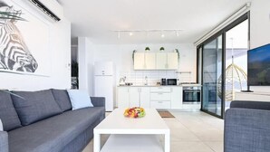 A modern living room with a gray sofa, a white coffee table with fruit, and a kitchen in the background with white cabinets and a large window.
