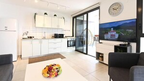 A modern kitchen with white cabinets, a dining area with a fruit bowl, and a large flat-screen TV mounted on the wall.