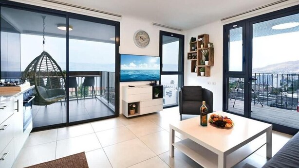 A modern living room with a large TV, a dining table with a bottle and fruit, a black chair, and a view of the city through large windows.