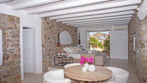 A cozy room with stone walls, a wooden ceiling, a round table with white chairs, a sofa, and a view of a hillside. - Topos exclusive Mykonos-villa Kymothoe (Agios Ioannis Diakoftis)