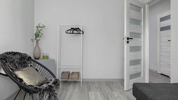 Una stanza con una parete bianca, una poltrona in vimini nero con cuscino, una scaffalatura bianca con cesti e una porta con finitura in legno bianco.