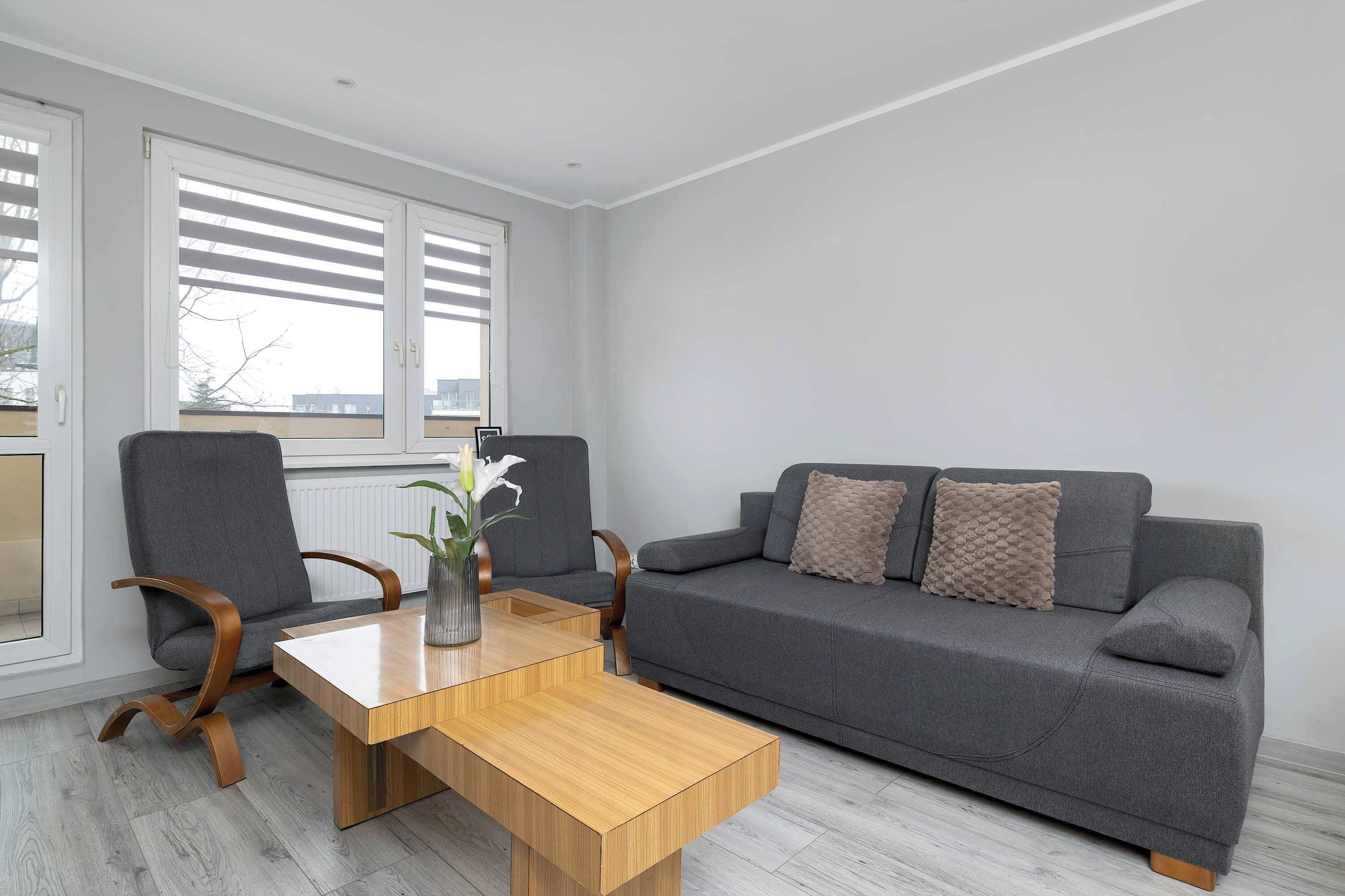 A modern living room with a grey sofa, two armchairs, a wooden coffee table, and a large window with blinds.