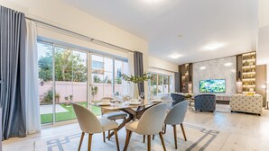 A modern living room with a dining area, large windows, and a flat-screen TV. - 4BR + Maid’s Room Villa Near Nad Al Sheba Mall, With Easy Access to Major Dubai Attractions (Dubai)