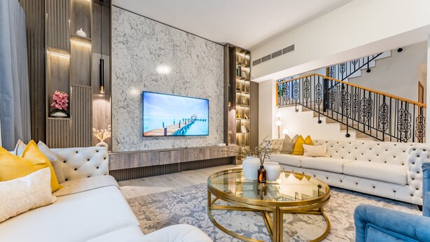 A modern living room with a large flat-screen TV, a white sofa, a round coffee table, and a staircase with black railings. - 4BR + Maid’s Room Villa Near Nad Al Sheba Mall, With Easy Access to Major Dubai Attractions (Dubai)