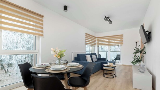 A modern living room with a dining area, large windows with blinds, a sofa, and a TV mounted on the wall.