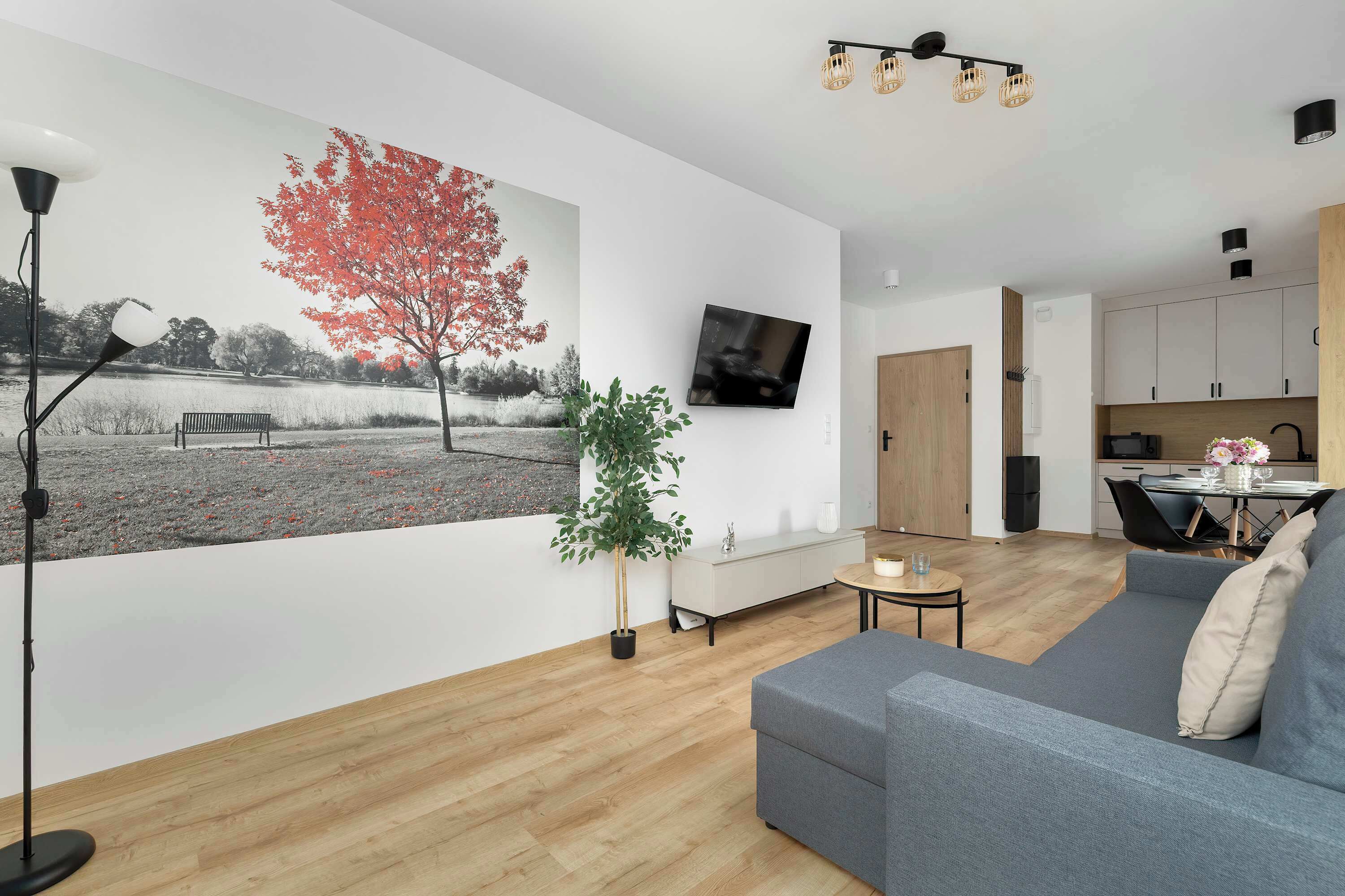 A modern living room with a large wall mural, a grey sofa, a small table, a floor lamp, and a kitchen area in the background.