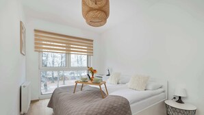A modern bedroom with a large window, a bed with a grey quilt, a bedside table with a lamp, and a radiator.