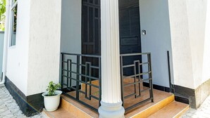 A building with a white facade, a balcony with black railings, and a potted plant on the ground floor. - Tadre House's (Arusha)