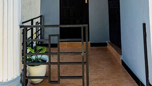 A hallway with a tiled floor, a potted plant, a metal railing, and a door with a black shutter. - Tadre House's (Arusha)