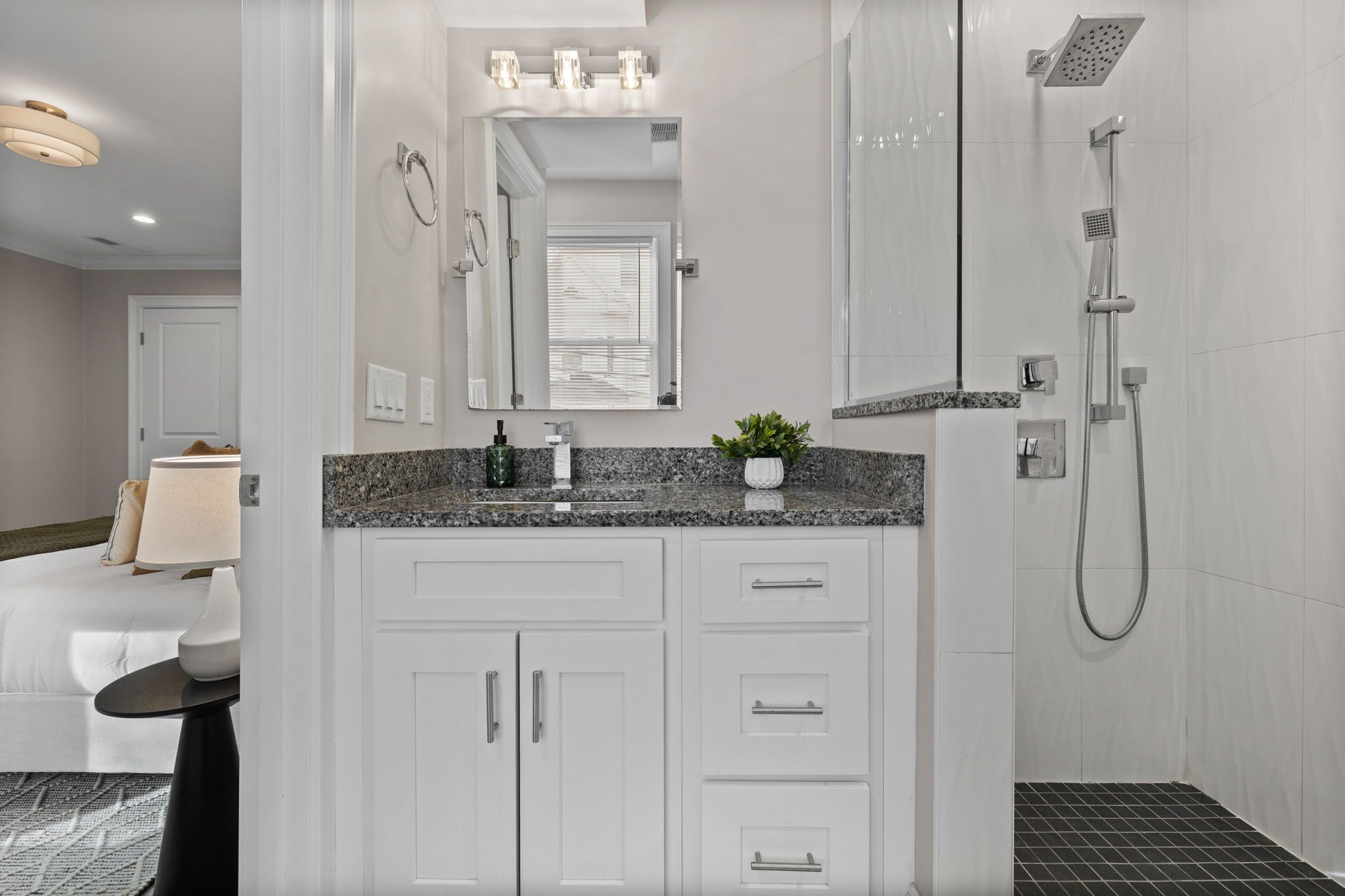 A modern bathroom with white cabinetry, a granite countertop, a large mirror, a shower with a handheld showerhead, and a potted plant on the counter.