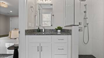 A modern bathroom with white cabinetry, a granite countertop, a large mirror, a shower with a handheld showerhead, and a potted plant on the counter.