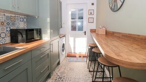 A compact kitchen with a microwave, washing machine, and a wooden bar area with stools. - The Corner Cottage (Wotton-under-Edge)