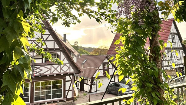 A traditional half-timbered building with a balcony, surrounded by lush greenery and purple flowers.