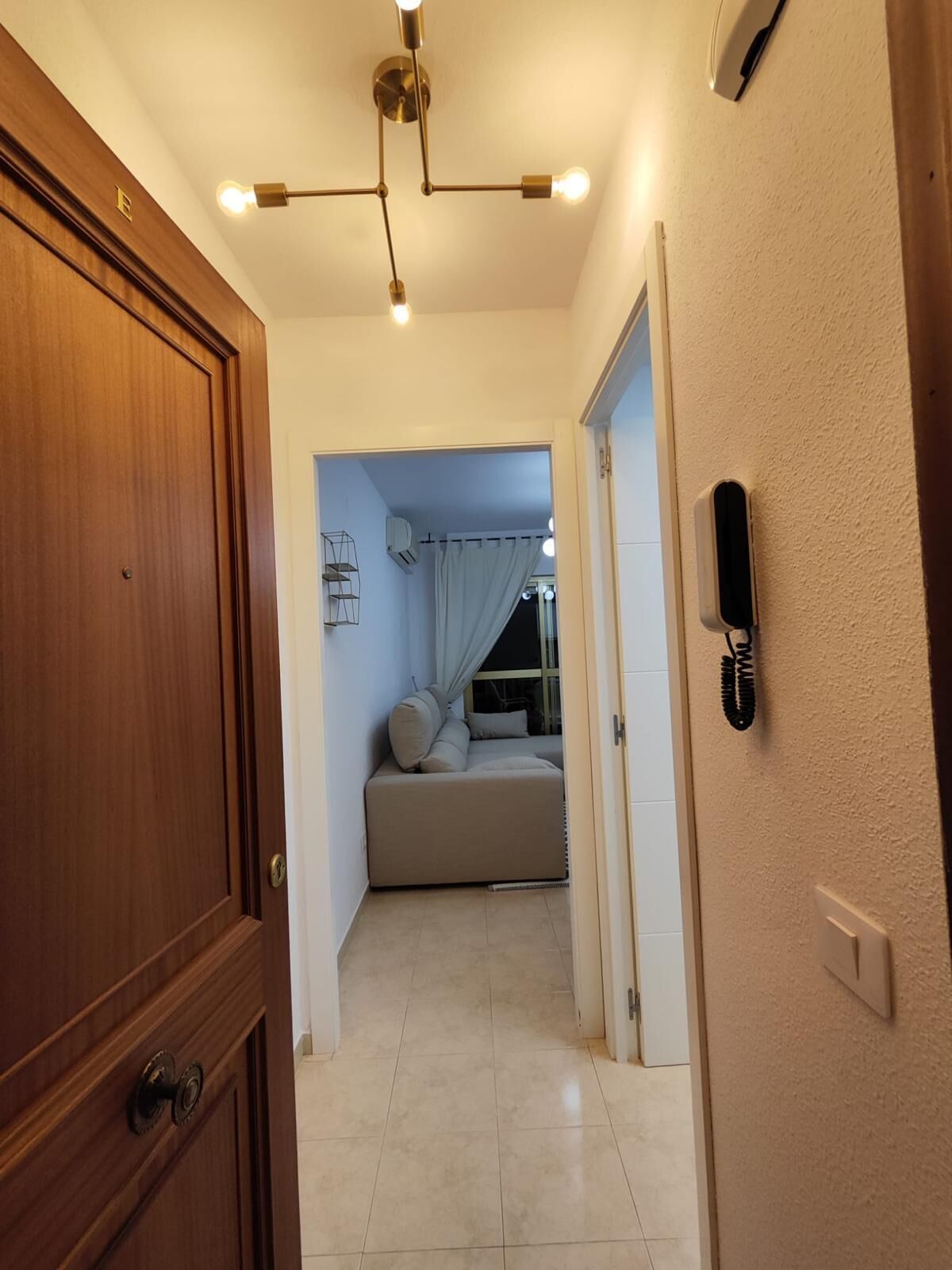 A hallway leading to a bedroom with a bed, a wooden door, a ceiling light fixture, and a wall-mounted phone.