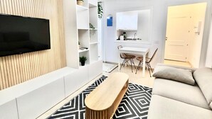 Un salon moderne doté d’un téléviseur à écran plat, d’un banc en bois, d’un tapis à motifs, d’un canapé et d’un coin repas avec une table et des chaises blanches.