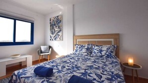 Een slaapkamer met een bed, een stoel, een nachtkastje, een kunstwerk aan de muur en een raam.