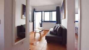Een woonkamer met een bank, een salontafel en een televisie. Er is een raam met gordijnen en een deur naar een andere kamer.