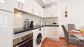 A modern kitchen with white cabinets, a built-in oven, a washing machine, and a dining area with a glass table and chairs. - Book Jet - NinoMar Heart Morro Jable (Morro Jable)