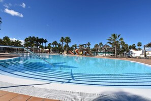 Ein klarer, blauer Swimmingpool mit einer von Palmen gesäumten Liegefläche, einer Wasserrutsche und einer Struktur mit einem Reetdach.