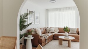 Un salon moderne avec un canapé en cuir marron, deux petites tables d’appoint blanches, un fauteuil en osier et une grande plante.
