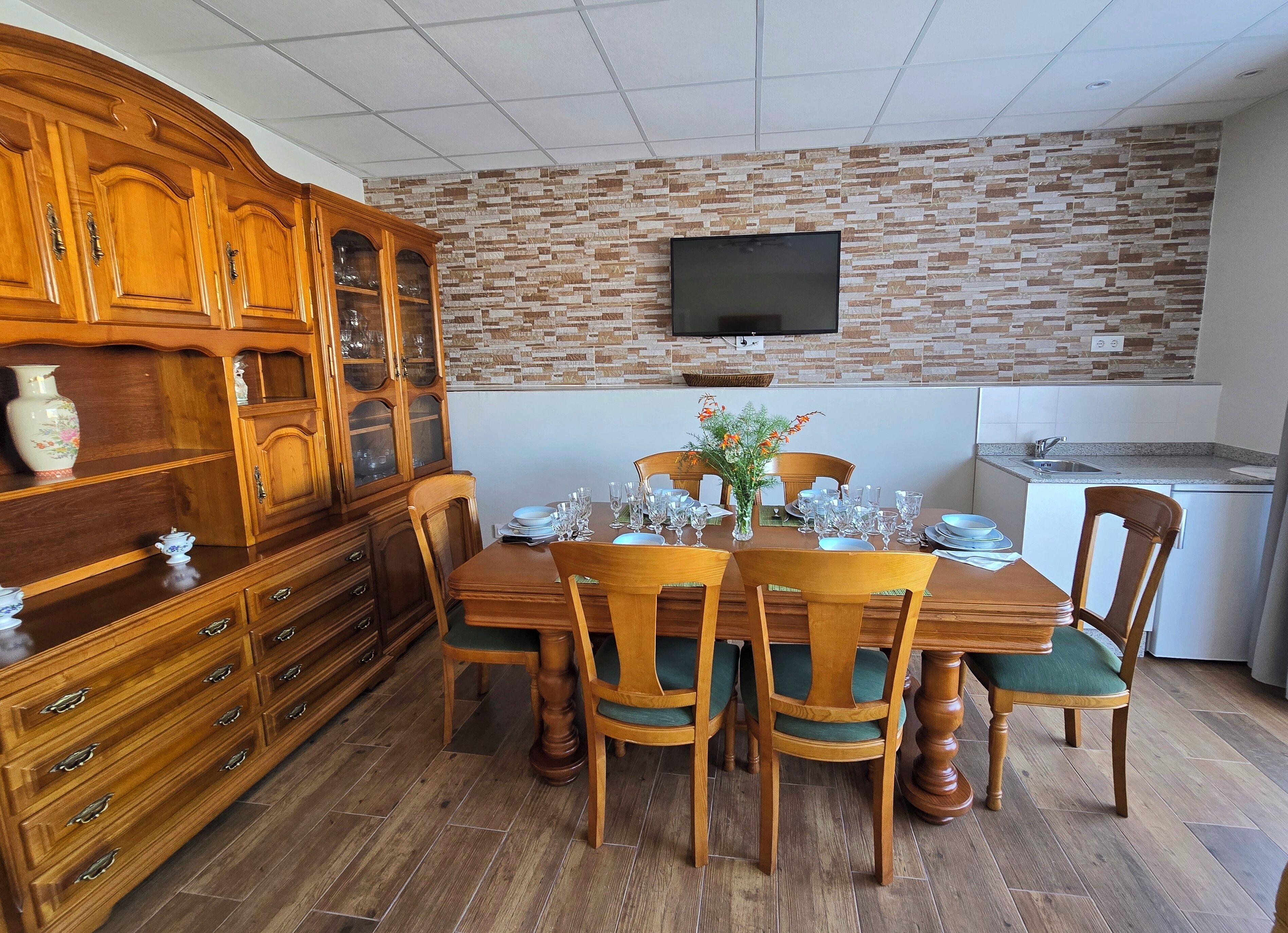 Uma área de refeições com uma mesa de madeira posta para quatro pessoas, rodeada por cadeiras com almofadas verdes. Um armário de madeira com portas de vidro está à esquerda, e uma TV de tela plana está fixada na parede acima da mesa.