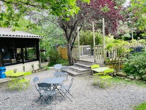 A covered outdoor seating area with tables and chairs, surrounded by greenery and a wooden deck.