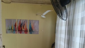 A room with a wooden floor, a bench, a guitar, a ceiling fan, and three paintings of sailboats. - Turtle Garden Lodge  (Mabul Island)