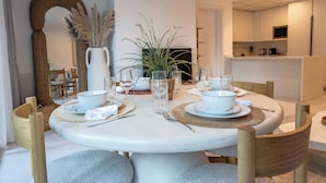 Un comedor con una mesa redonda dispuesta para cuatro personas, rodeada de sillas de madera. Hay dos jarras blancas, un florero de vidrio con una planta y un mantel tejido en la mesa.