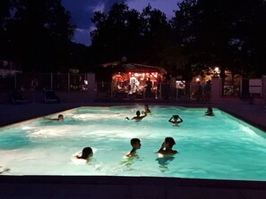 Une piscine où des personnes se baignent et s’amusent, entourée d’arbres et d’un bâtiment dont les lumières rouges se détachent à l’arrière-plan.