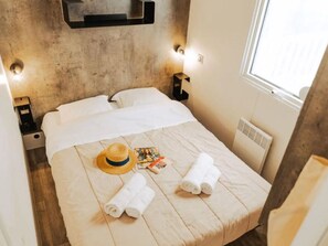A bed with white linens, a straw hat, rolled towels, and a magazine on a wooden floor. - Spacious 40m² Mobile Home with Terrace (Saint-Jean-de-Monts)