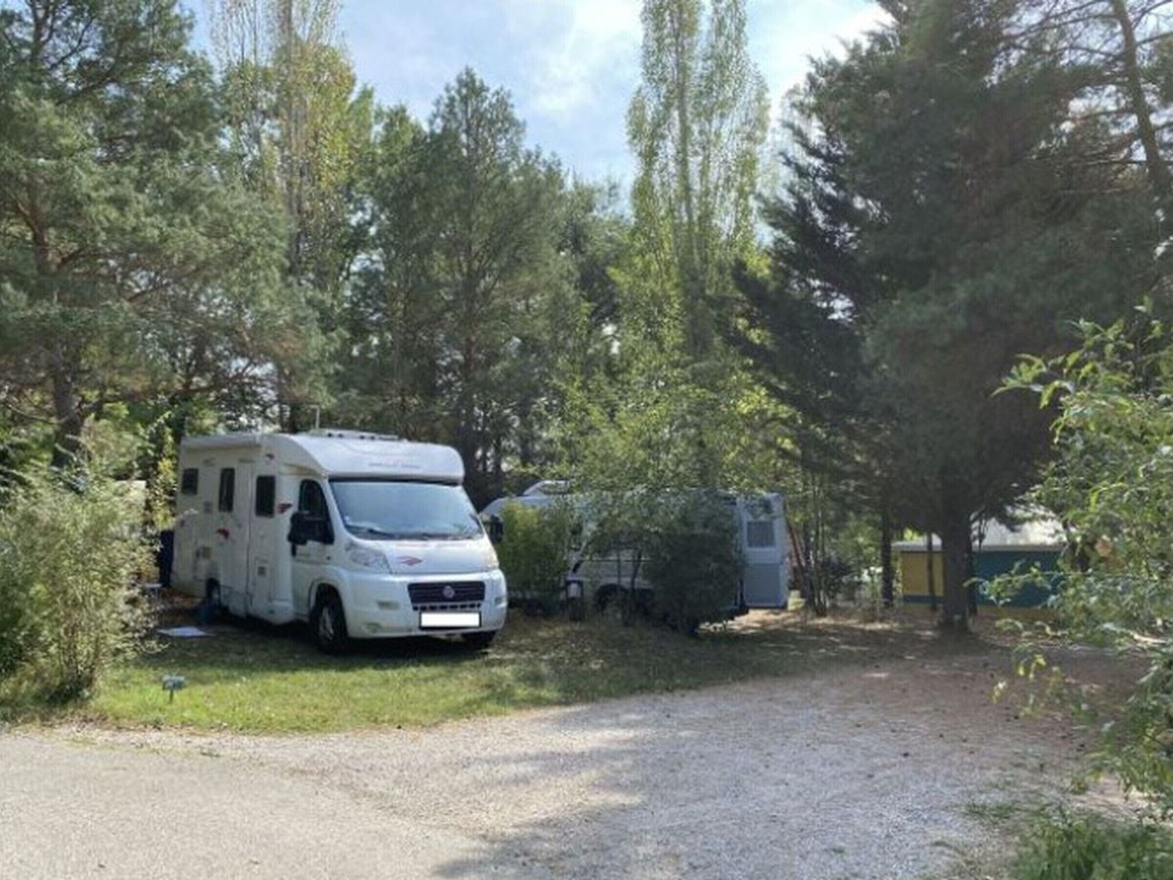 Un camping-car blanc est garé dans une zone boisée, à proximité d’un chemin de gravier.