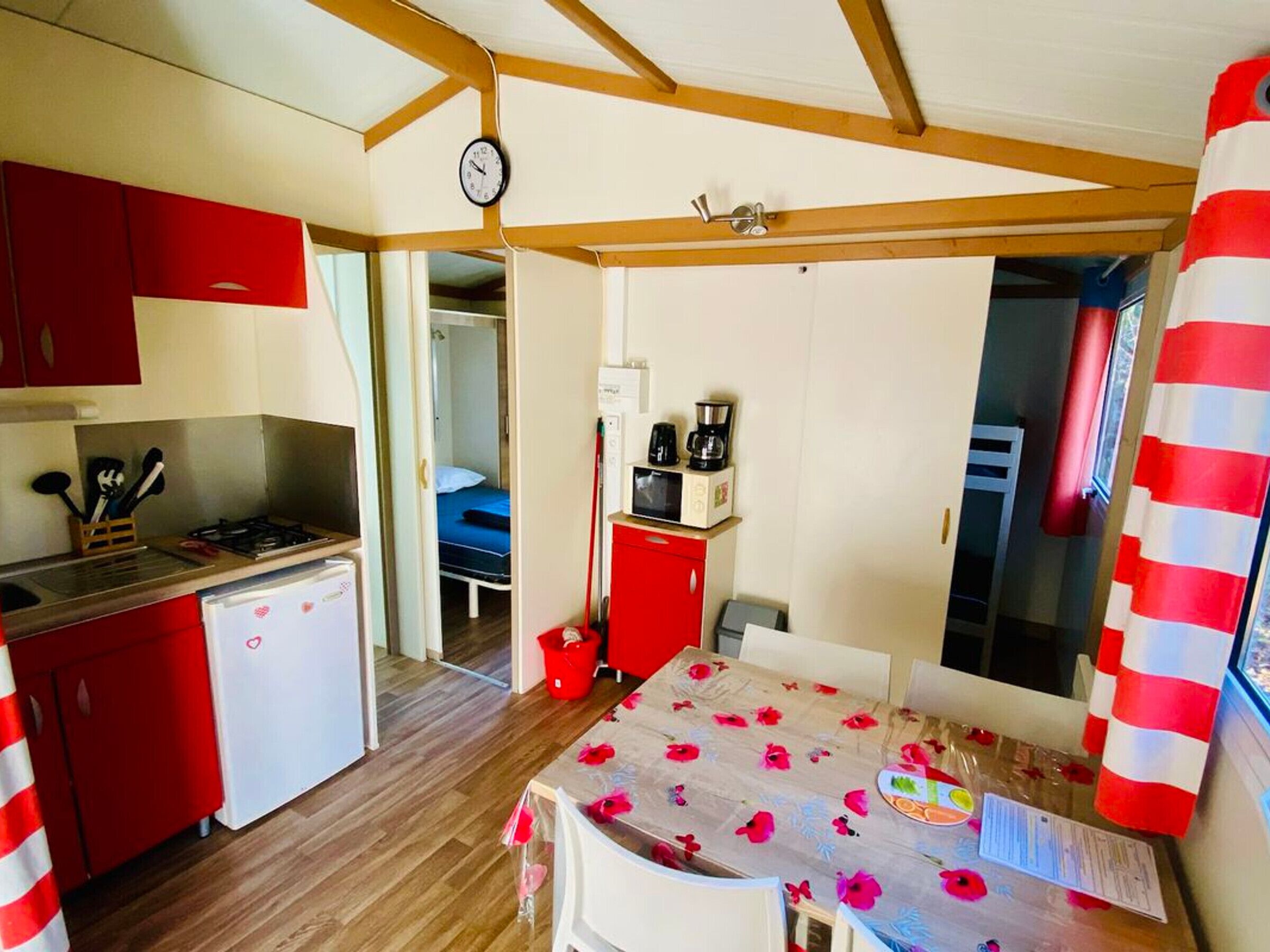 Una habitación compacta con kitchenette, una cama, una mesa de comedor con mantel de flores y una cortina a rayas rojas.