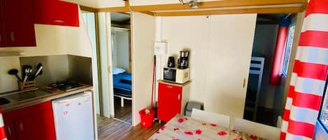 Una habitación compacta con kitchenette, una cama, una mesa de comedor con mantel de flores y una cortina a rayas rojas.